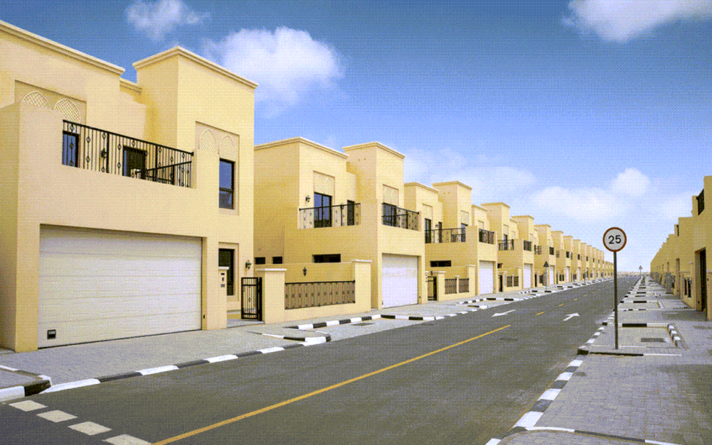 Nad Al Sheba Apartments area with modern buildings, an open road, and a blue sky dotted with clouds.